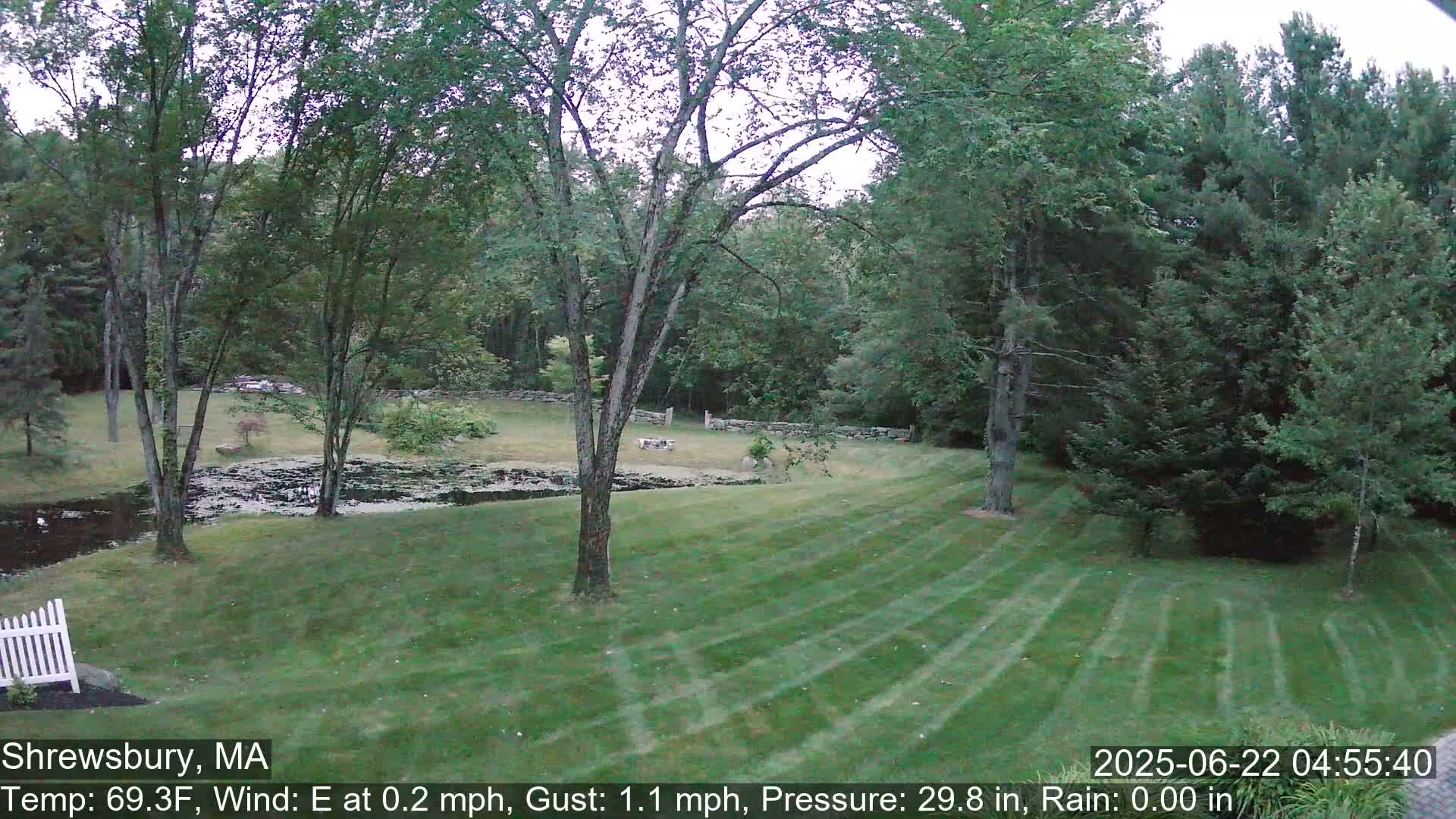 A neatly mown lawn slopes down to a small pond, bordered by trees and a stone wall, under a mostly cloudy sky.