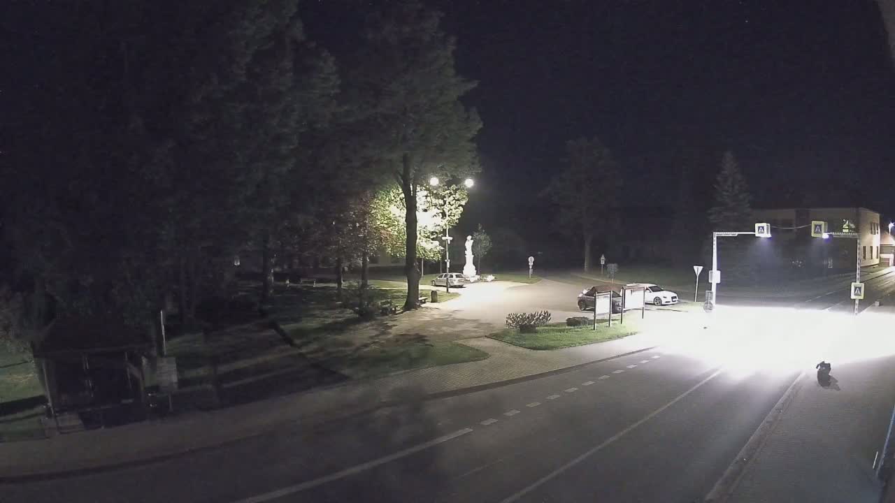 A nighttime scene shows a small town square with a statue, a few cars, and a street with traffic, all under dark skies.