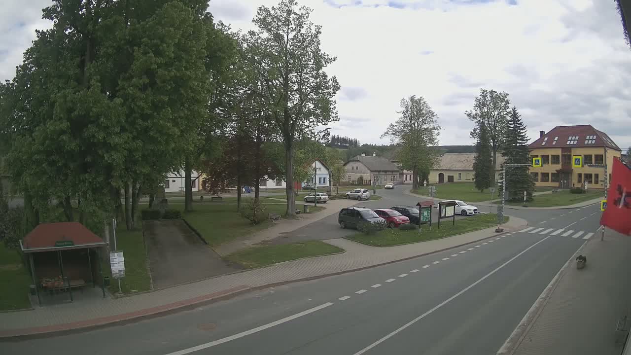 A partly cloudy day reveals a town square with several cars parked near buildings and a bus stop, all surrounded by lush green trees.