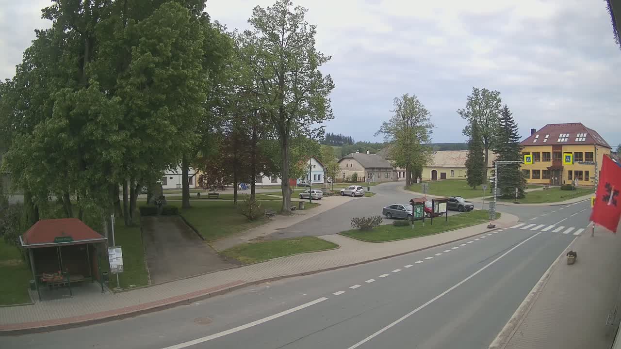 A town square with several cars parked, a bus stop, and a two-story building under a cloudy sky.
