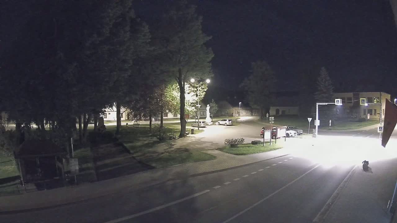 A nighttime view of a town square with a statue, several parked cars, and a street with a vehicle approaching, all surrounded by trees and buildings under dark skies.