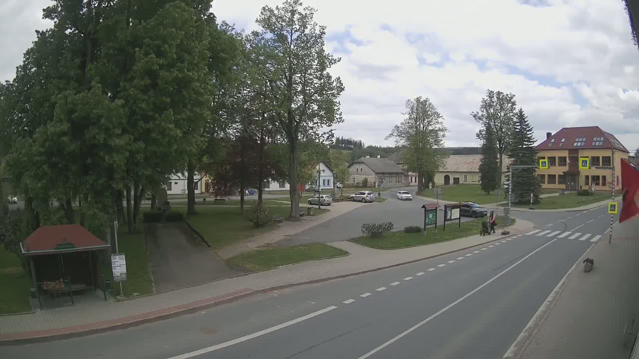 A partly cloudy day reveals a town square with several cars parked and a few pedestrians walking, surrounded by lush green trees and buildings.