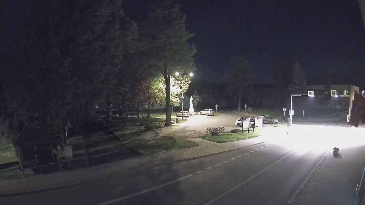 A nighttime view of a small town square with a few cars parked and a statue in the center, illuminated by streetlights.