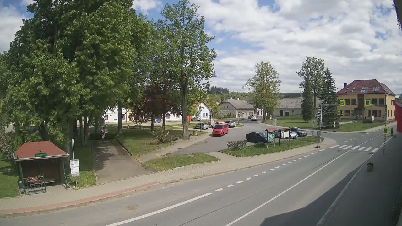 A village square with a few cars, lush green trees, and buildings under a partly cloudy sky.