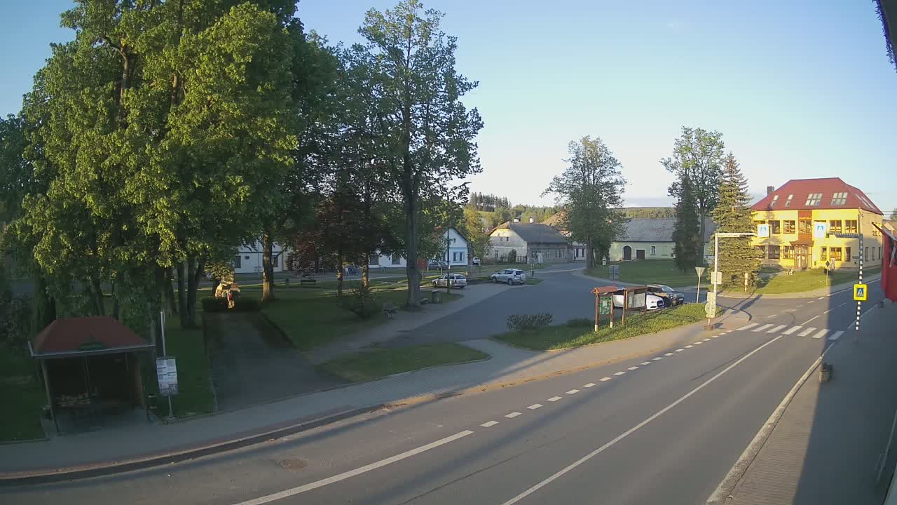 A sunny day in a small town square with several cars parked, green grass, and leafy trees surrounding buildings.