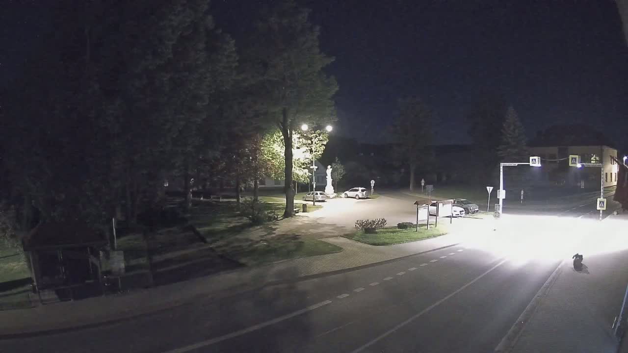 A nighttime view of a small town square with a statue, a few parked cars, and a road with a vehicle approaching.