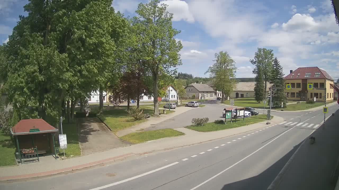 A sunny day in a small town square with a few cars parked, some buildings, and green trees surrounding a grassy area.