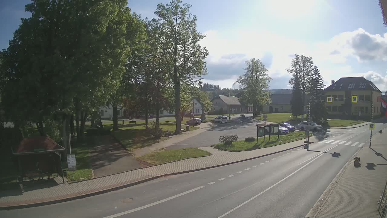 A sunny day reveals a village square with a few cars, surrounded by trees, buildings, and a paved road.