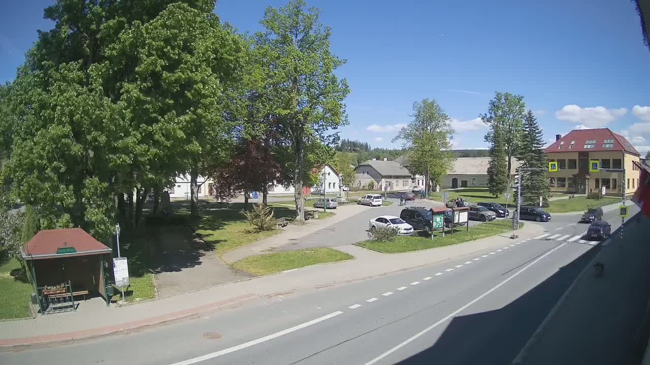 A sunny day in a small town square with several cars parked near a bus stop and several buildings in the background.