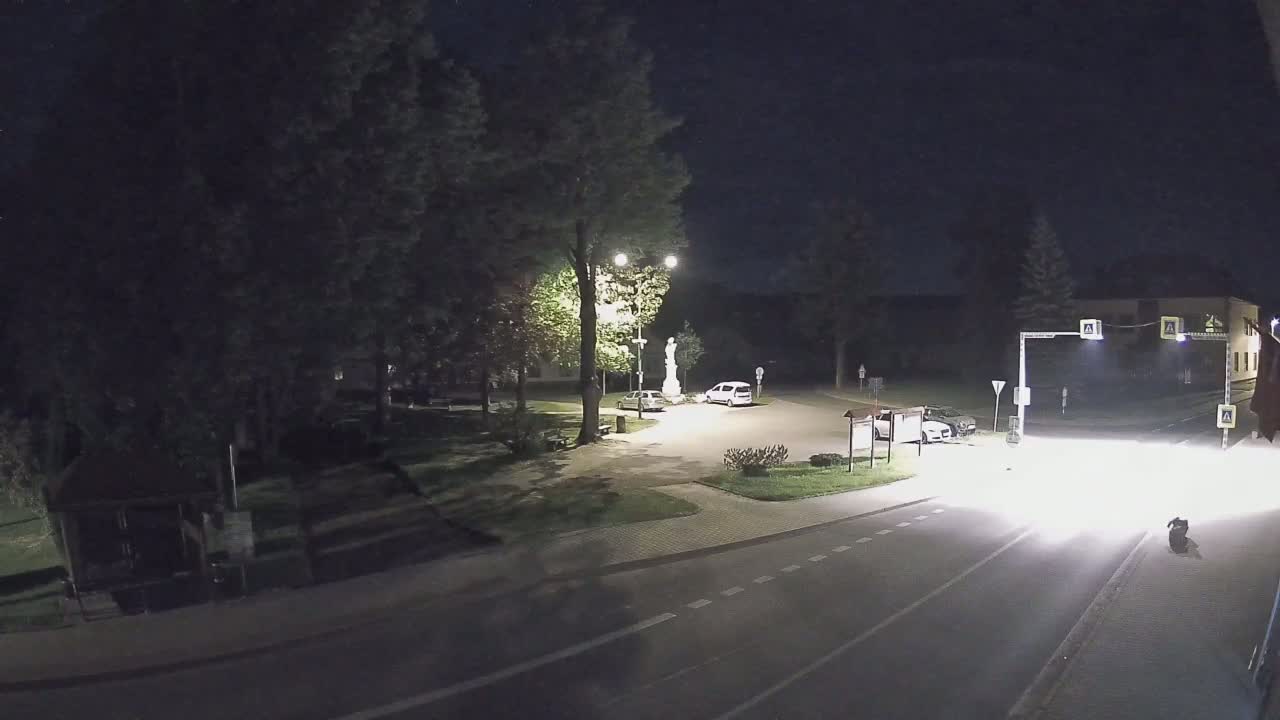 A nighttime scene shows a small town square with a statue, a few parked cars, and a road with a vehicle driving past.