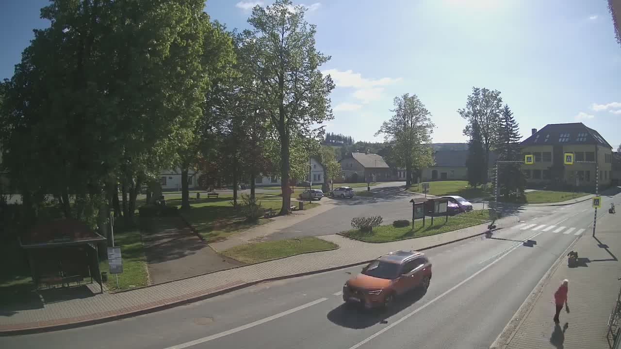 A sunny day reveals a small town square with several buildings, lush green trees, a pedestrian crossing, and an orange car driving on the road.