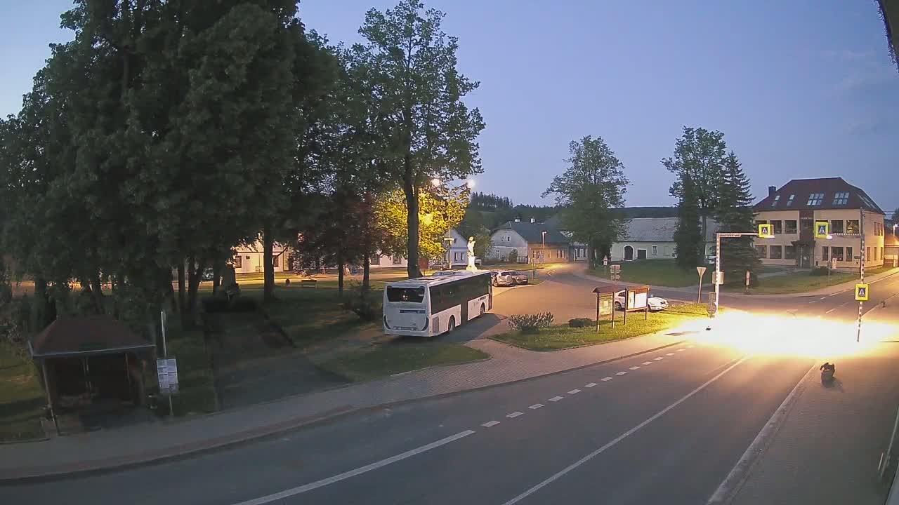 A bus is parked in a town square at dusk, near a road intersection with some cars and buildings visible under a clear sky.