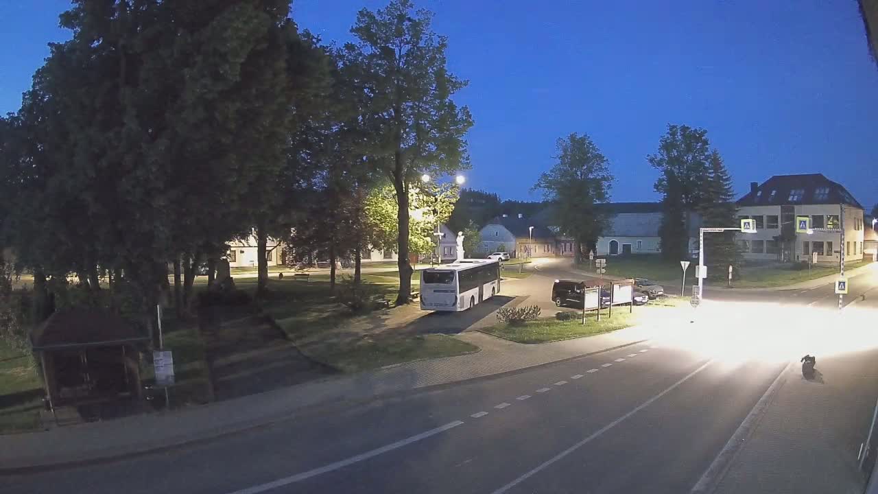 A white bus is parked in a village square at twilight, with cars parked nearby and a road visible in the foreground.