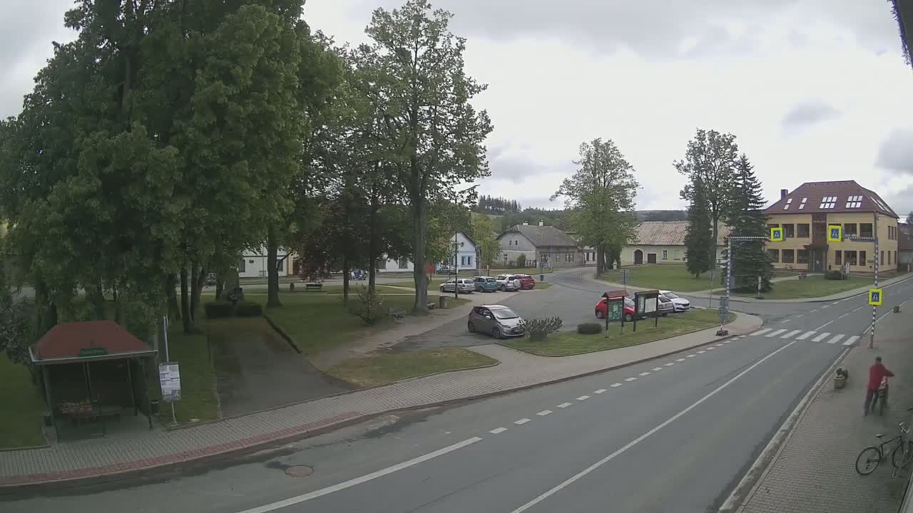 A small town square with several parked cars, a few buildings, green trees, and a mostly overcast sky.