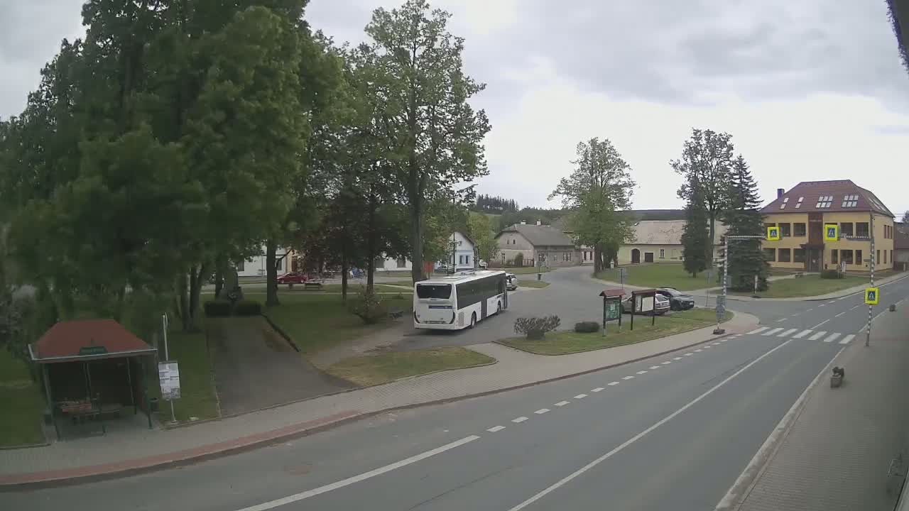 A white bus is parked in a village square under an overcast sky, near a two-story building and several cars.