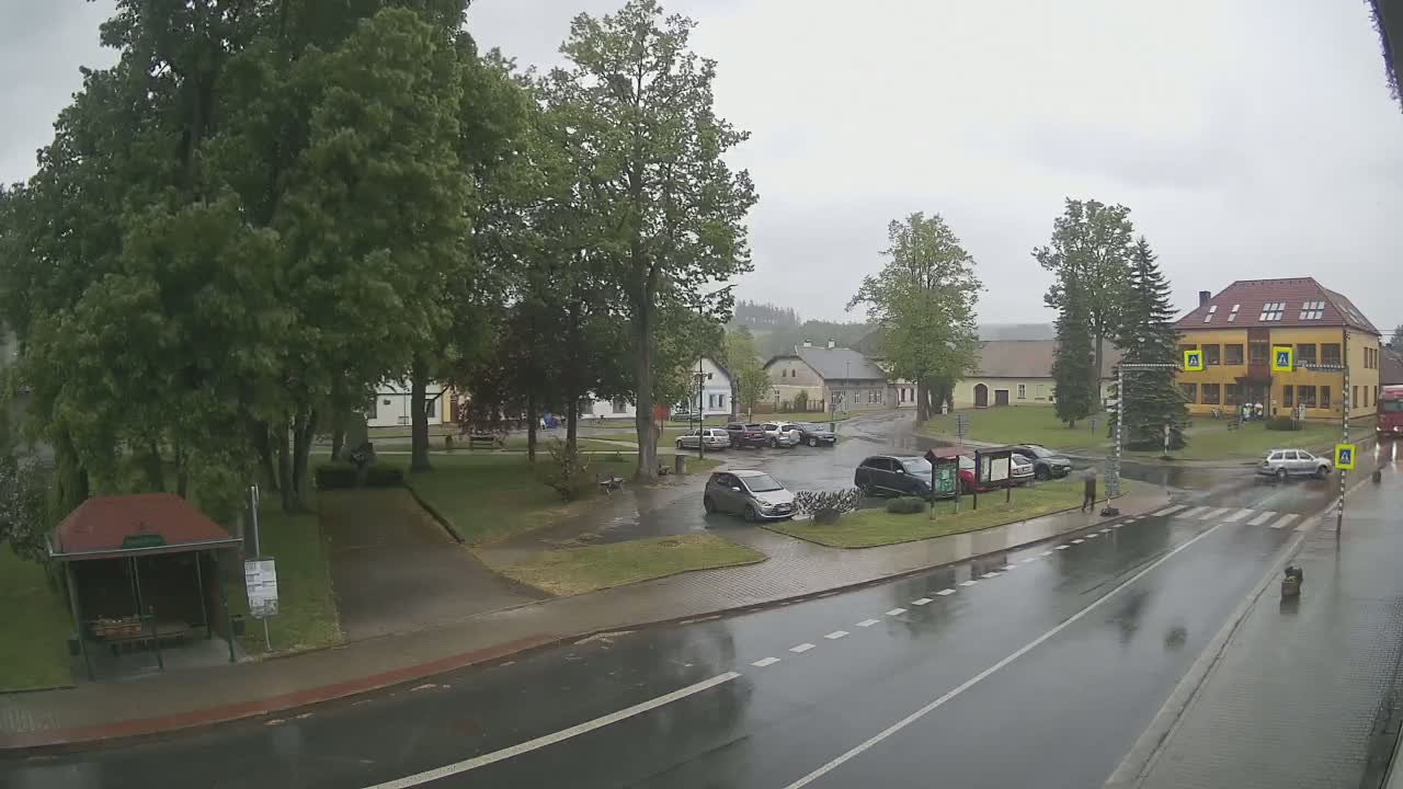A rain-soaked town square with several cars parked, a bus stop, and a two-story building in the background.