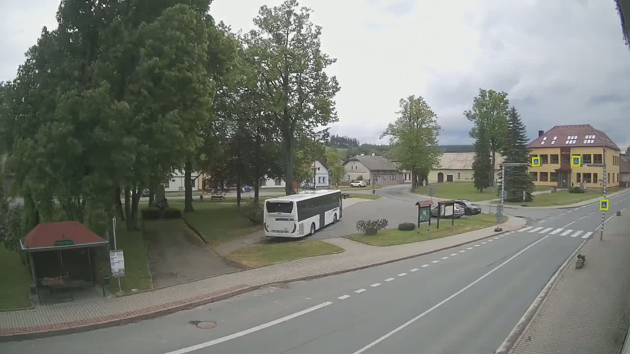 A white bus is parked in a village square under a partly cloudy sky, surrounded by buildings, trees, and a roadway.