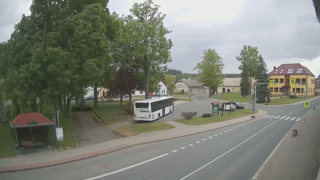 A white bus is parked in a village square on an overcast day, surrounded by buildings, trees, and a road.