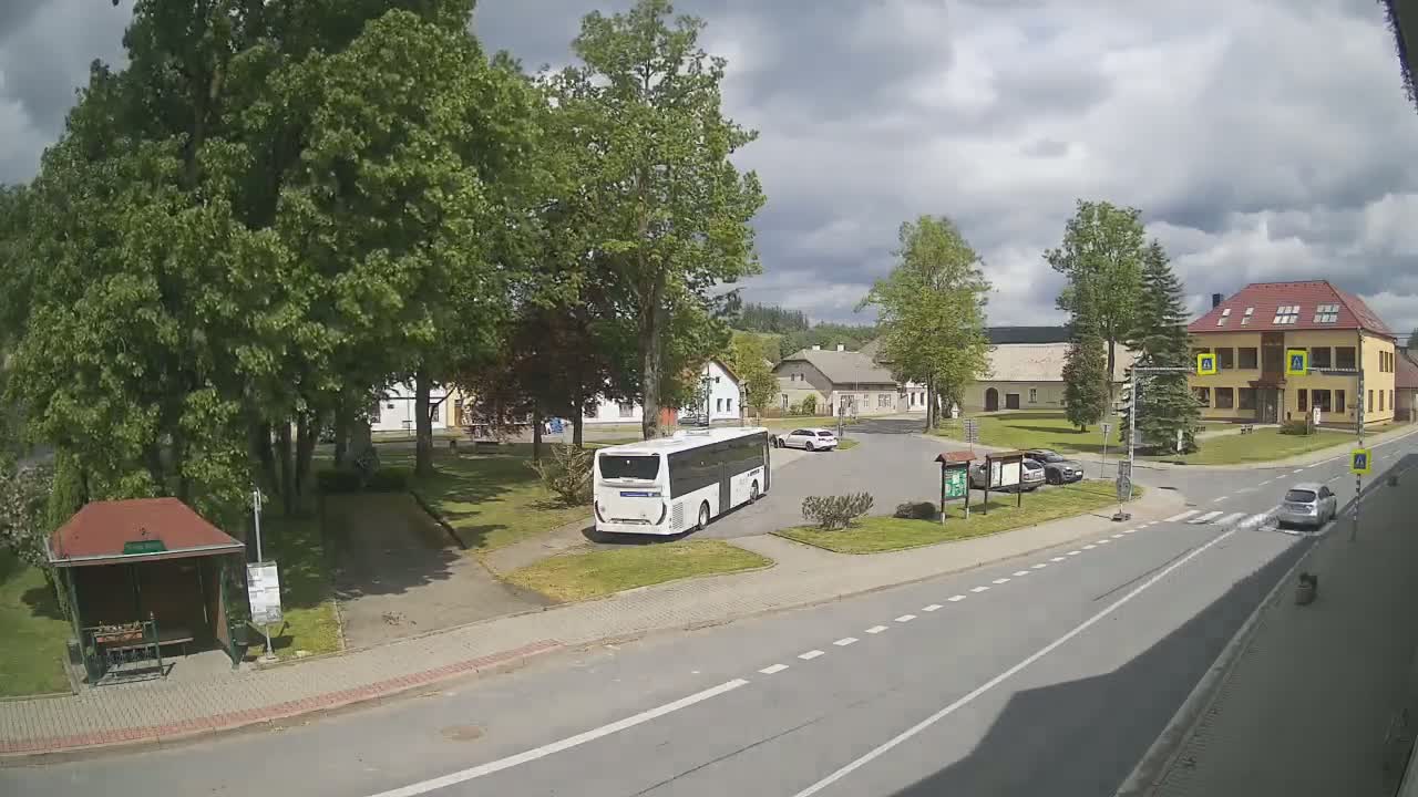 A white bus is parked in a village square under a partly cloudy sky, with several buildings and cars visible, near a bus stop.