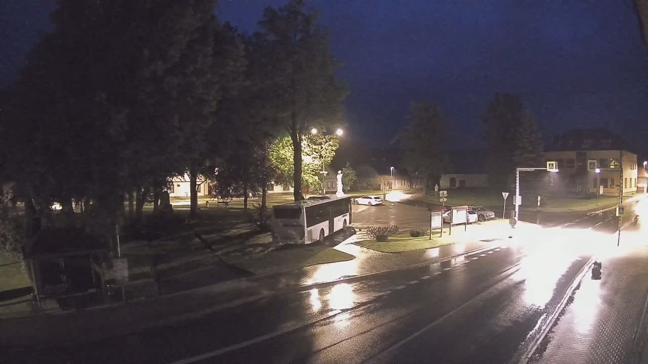 A bus is parked near a statue in a town square at night in the rain.