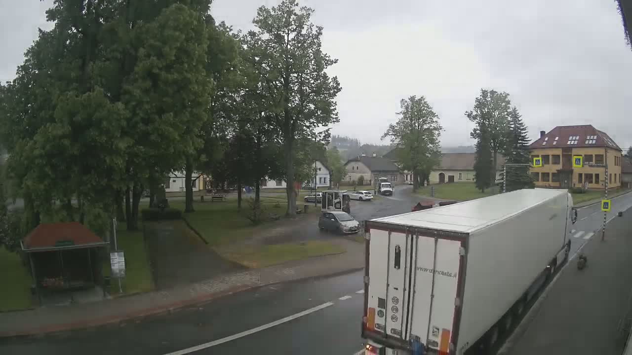 A rainy view of a town square with several cars, a large white truck, and buildings surrounded by lush green trees.