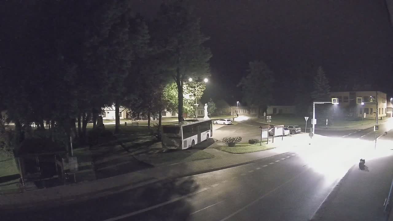 A nighttime view of a town square with a bus parked near a statue, a few cars, and trees surrounding the area under clear skies.