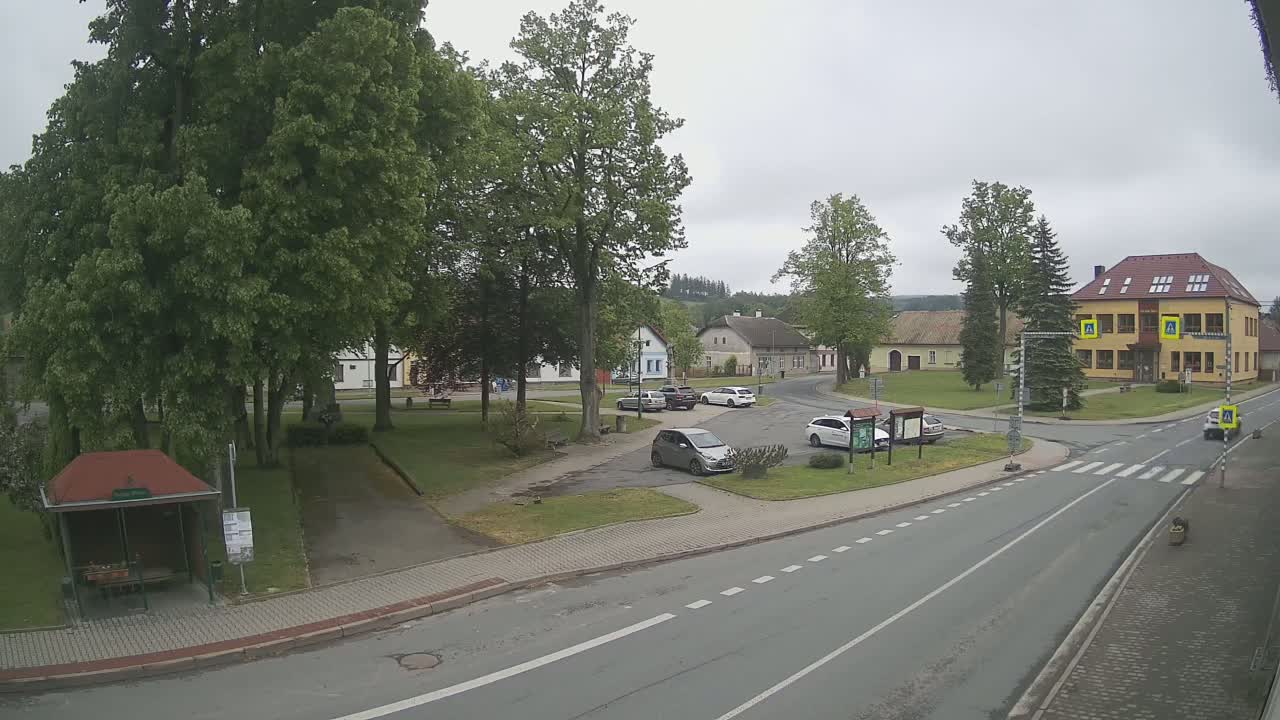 A small town square with several cars parked, surrounded by buildings and lush green trees under an overcast sky.