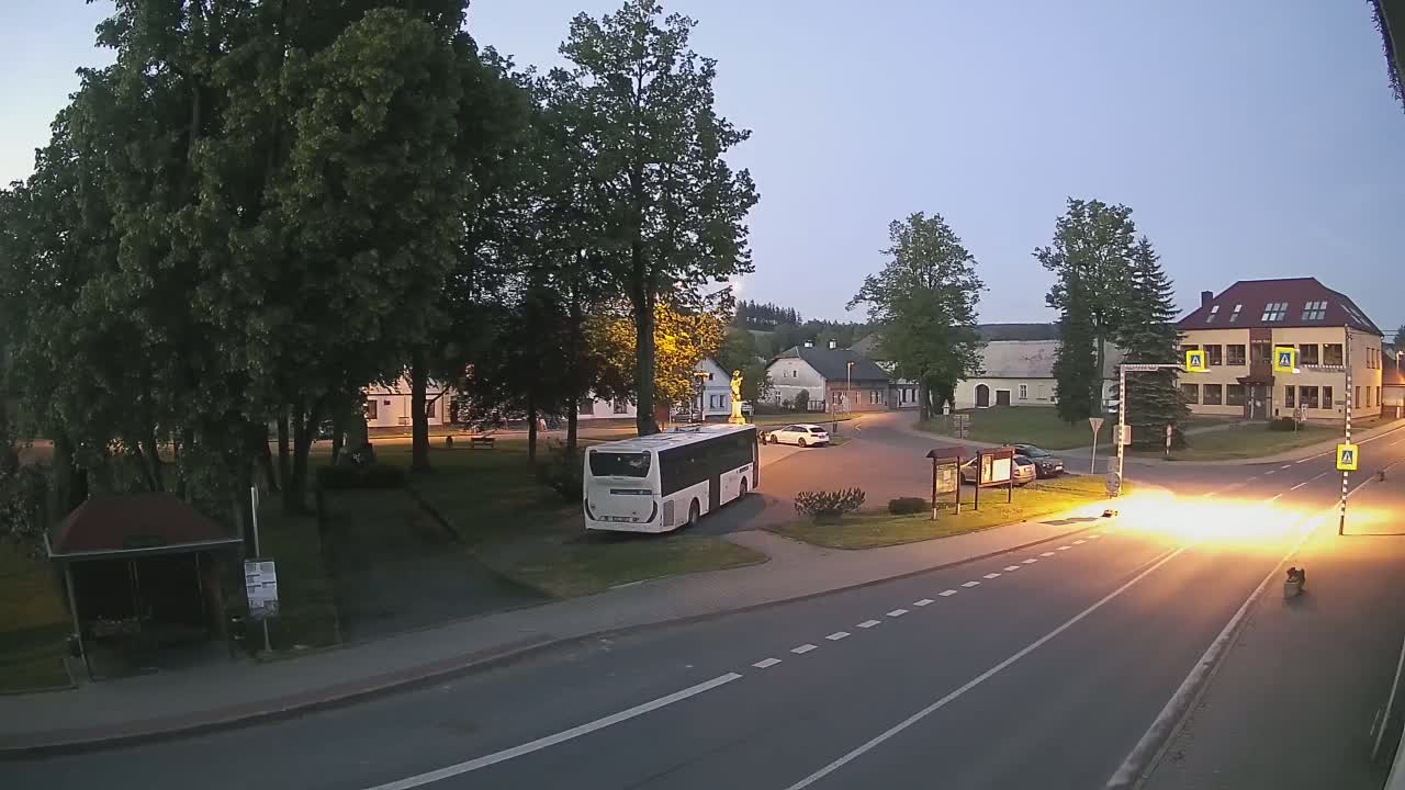 A white bus is parked in a village square at dusk, near a statue, some buildings, and a road with cars.