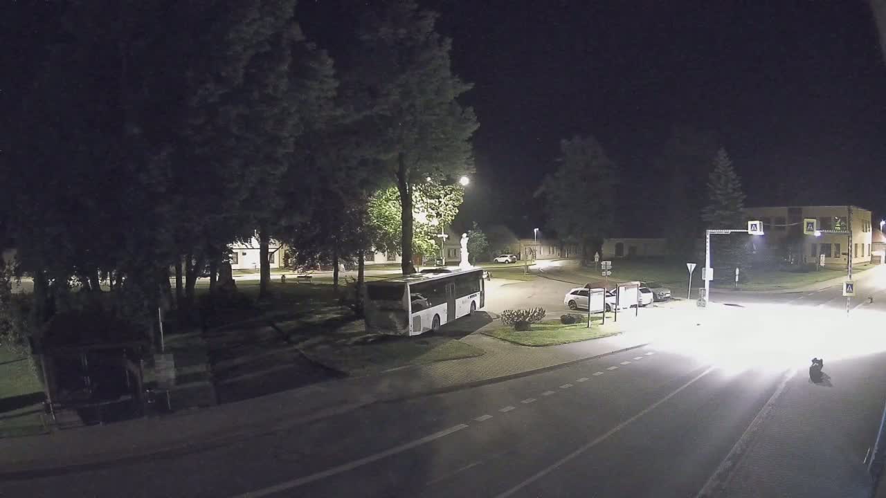 A nighttime scene shows a bus parked near a small town square with a few cars and a statue, under a clear, dark sky.