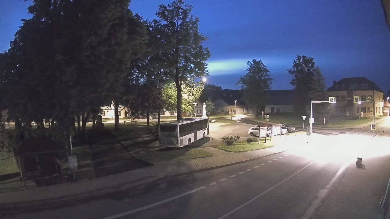 A nighttime view of a town square with a bus parked near trees and buildings, under a twilight sky.