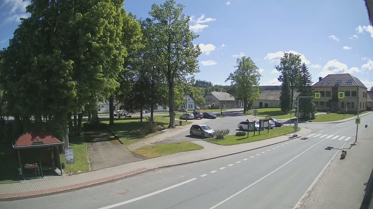 A sunny day reveals a village square with several cars parked, surrounded by lush green trees and buildings, a paved road passing through, and a bus stop nearby.