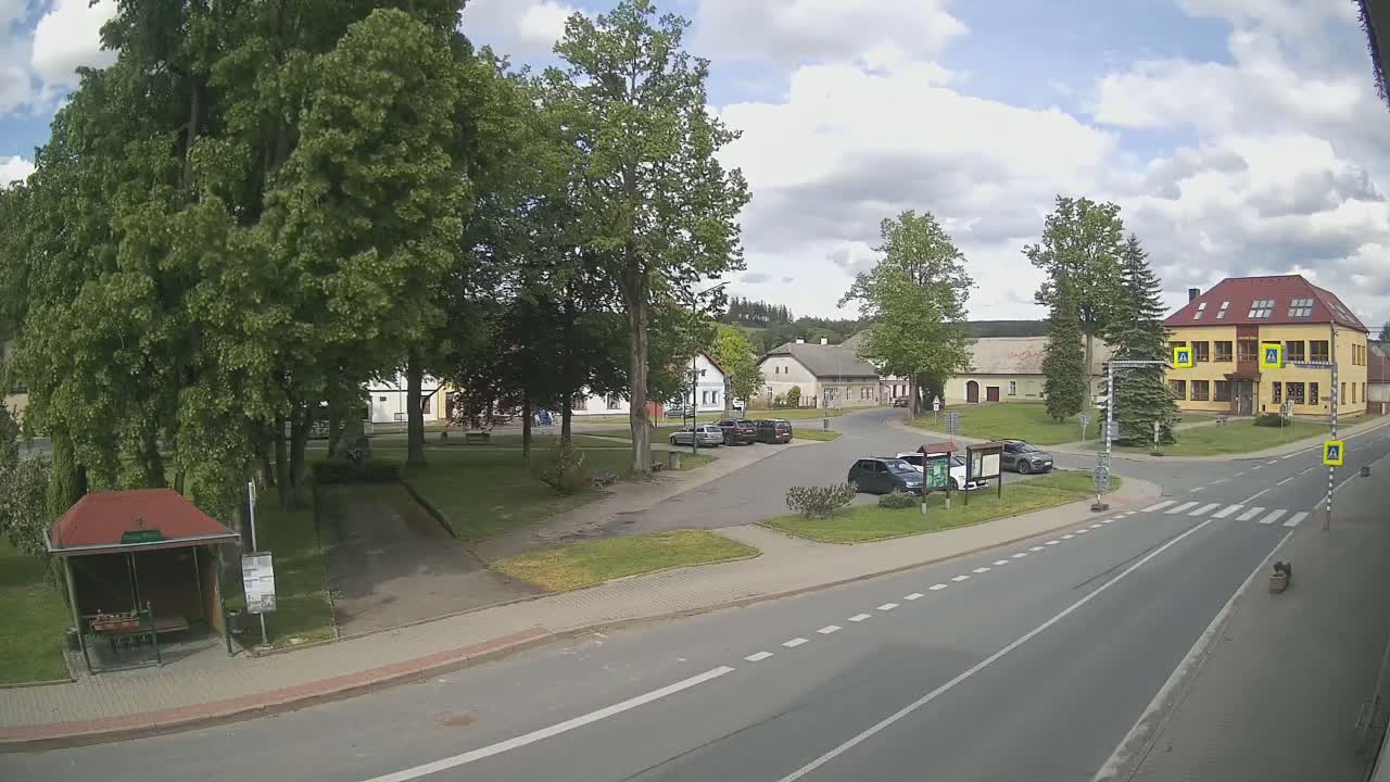 A partly cloudy day reveals a village square with several cars parked near a two-story building, a bus stop, and green spaces surrounded by leafy trees.
