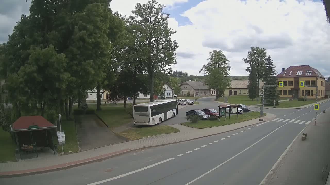 A partly cloudy day shows a town square with a bus stopped near a park and several cars parked in a lot, all near a crosswalk and road.