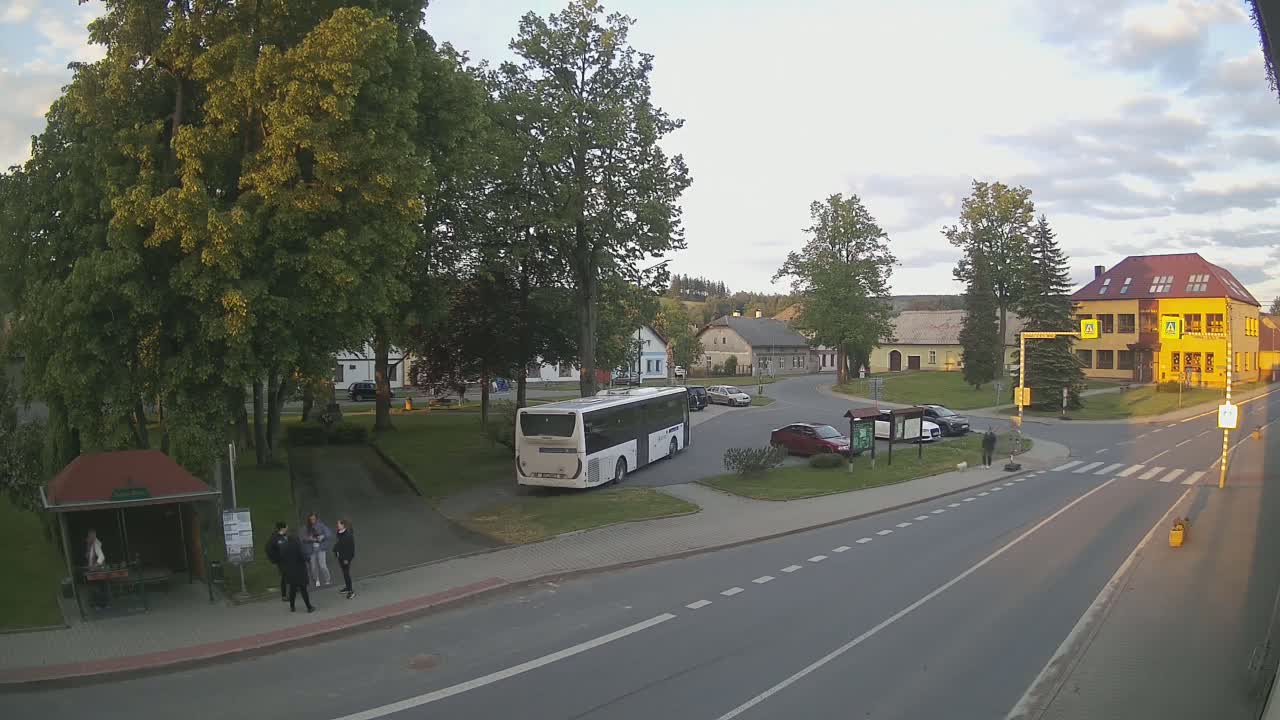 A bus is parked in a village square under a partly cloudy evening sky, near several cars, a bus stop, and some buildings.