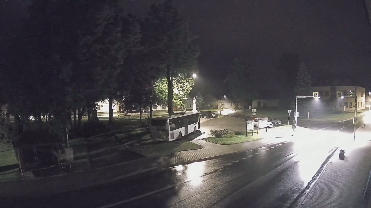 A nighttime view of a wet, empty town square with a bus parked near a statue, under dim streetlights.