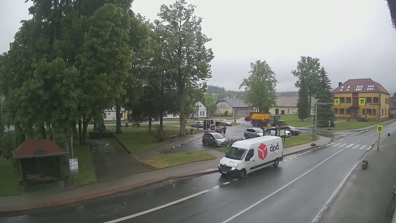 A wet, overcast town square with several cars and a delivery van, surrounded by green trees and buildings.