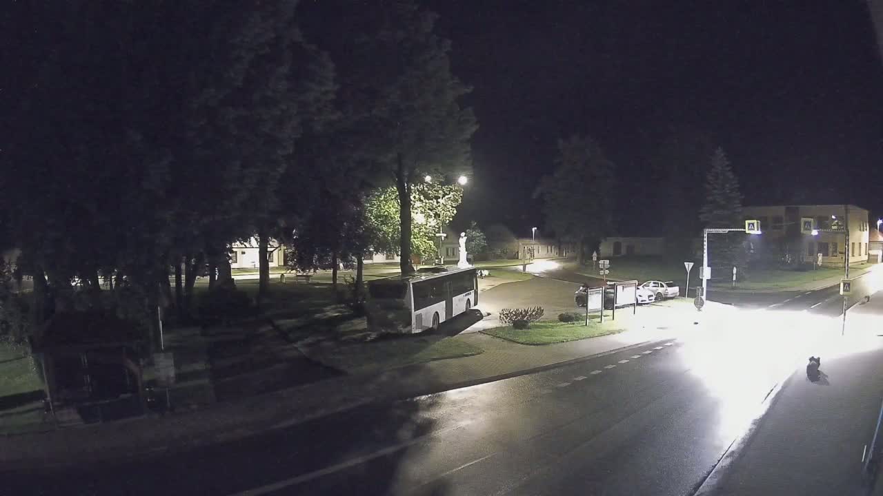 A nighttime view of a sparsely populated town square with a bus parked near a statue, a few cars, and wet roads under streetlights.