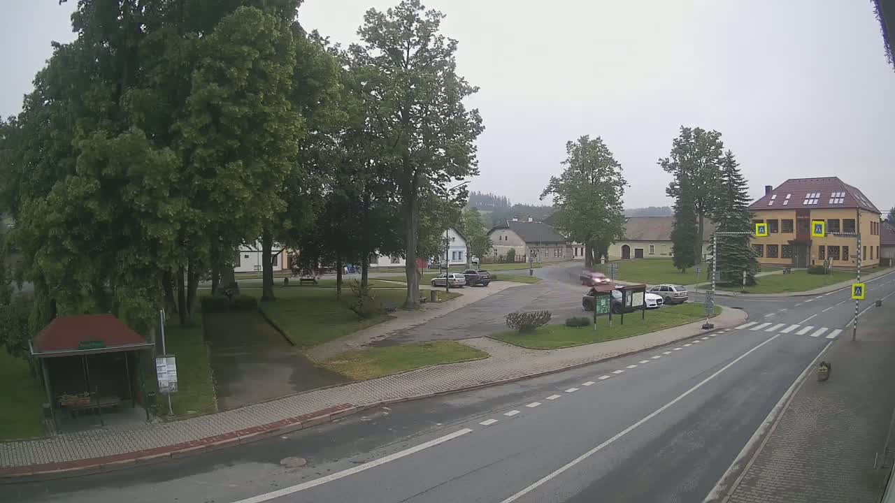 A village square with several cars, a bus stop, and buildings under an overcast sky.