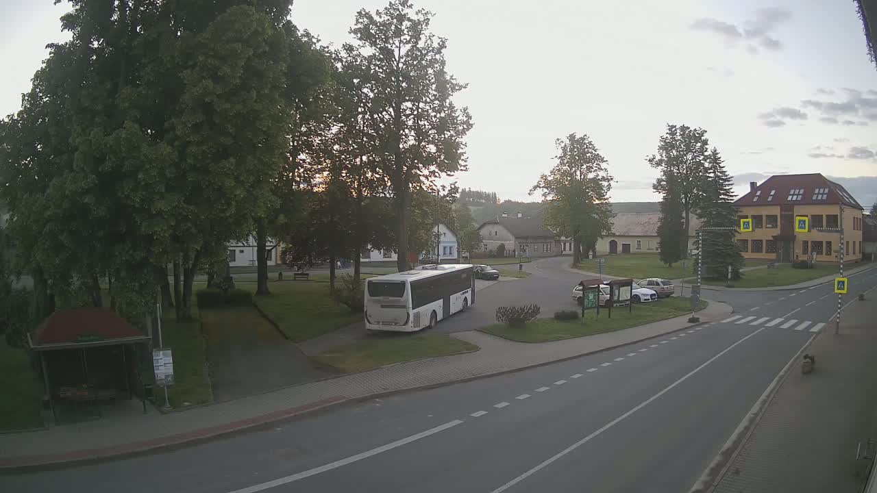 A white bus is parked in a village square under a mostly cloudy sky, near several buildings and leafy trees.