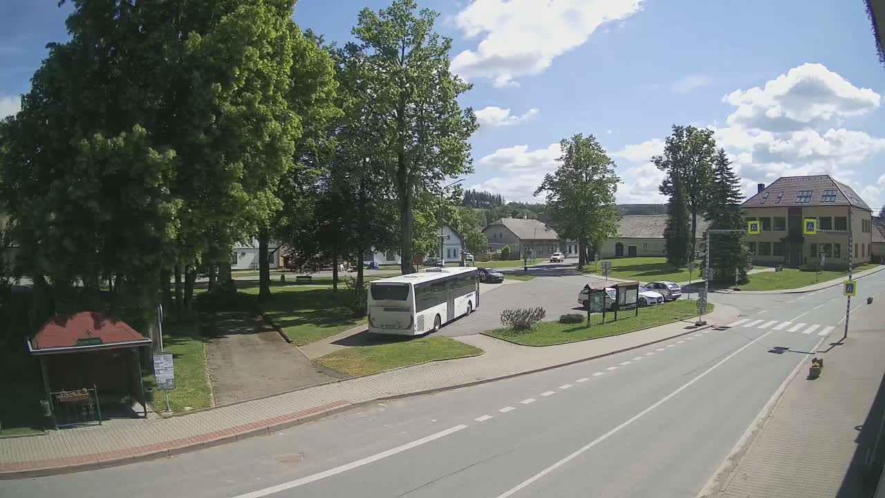 A white bus is parked in a village square on a sunny day with partly cloudy skies, surrounded by buildings, trees, and cars.