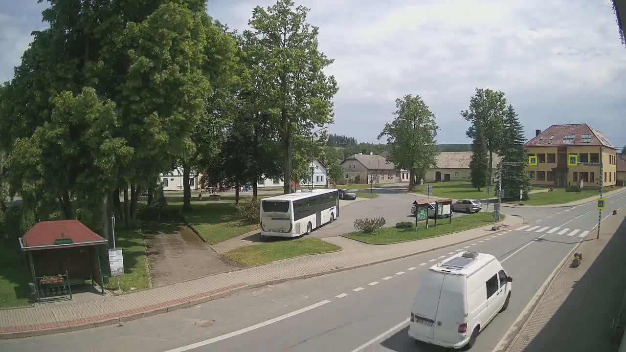 A partly cloudy day shows a village square with a bus and van, a small park, and several buildings, including a two-story house.