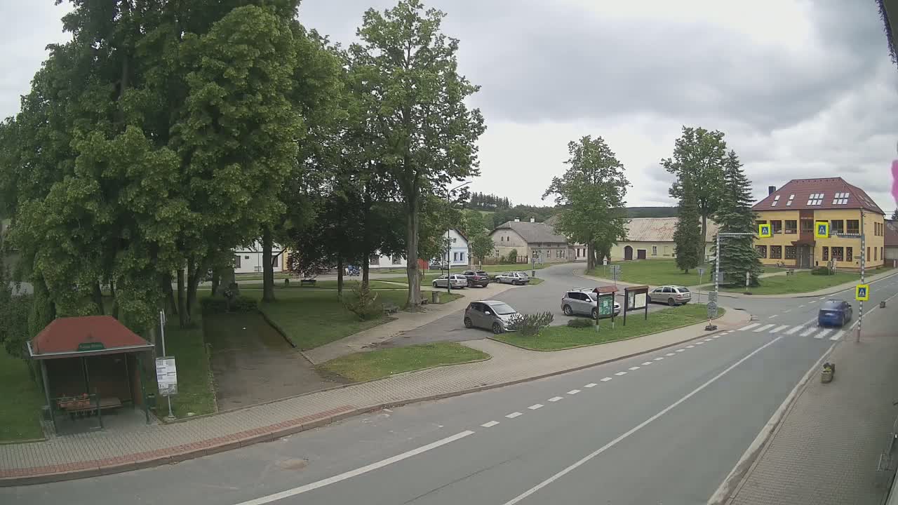 A mostly sunny, overcast day in a small town square with several cars parked and one driving, surrounded by lush green trees and buildings.
