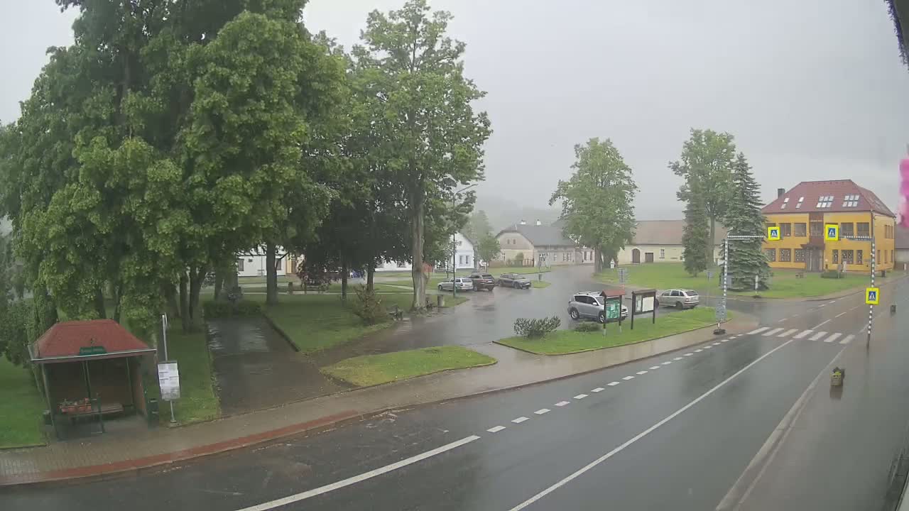 A rainy scene shows a village square with several cars parked, a bus stop, and buildings under a gray sky.