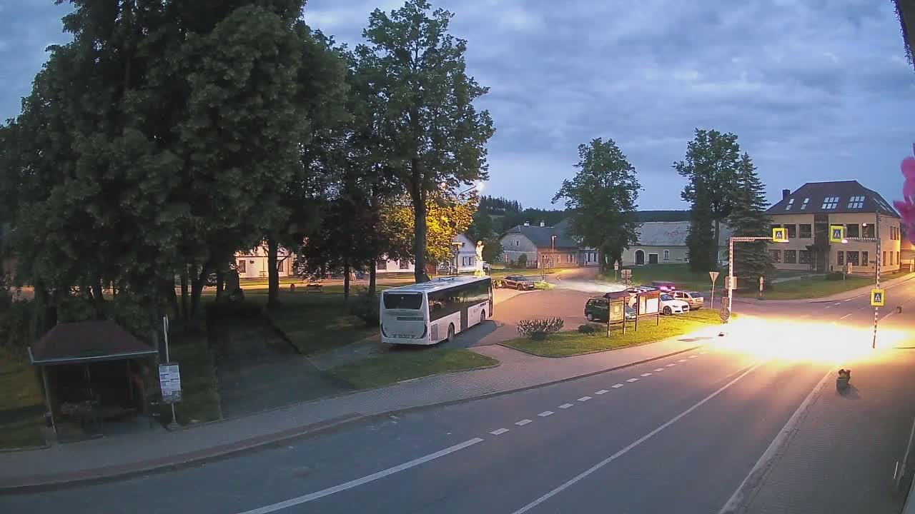 A bus is parked in a town square at dusk, with several cars at an intersection where a bright light is emanating from the road.