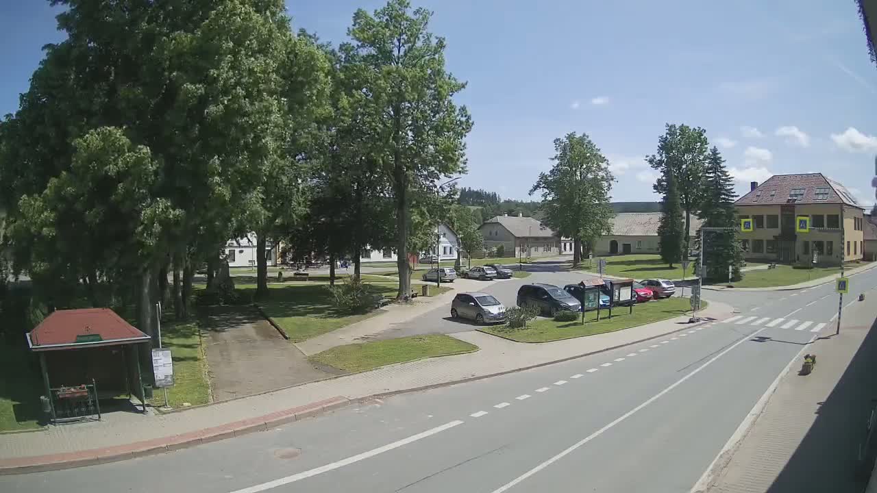 A sunny day in a village square with several cars parked near a grassy area and buildings surrounding it, a road with a pedestrian crossing is in the foreground.
