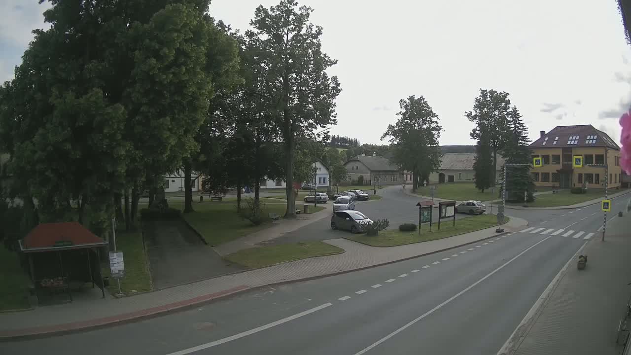 A mostly overcast day reveals a village square with several cars parked, a few buildings, and lush green trees surrounding a paved road.