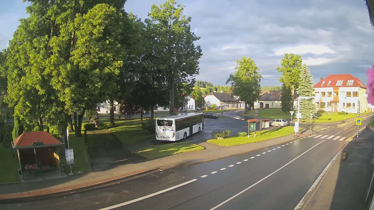 A white bus is parked in a small town square under a partly cloudy sky, with a wet road and lush green trees surrounding a building.