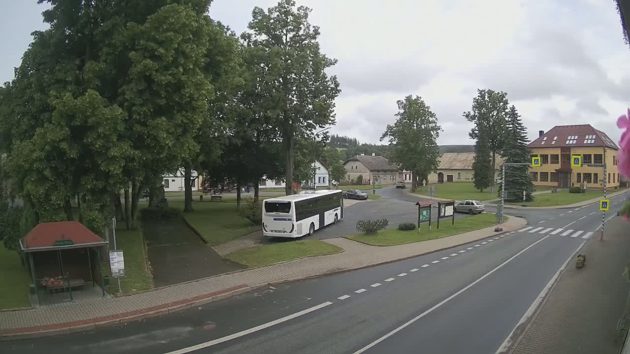A white bus is parked in a village square under an overcast sky, near several buildings, trees, and a small park.