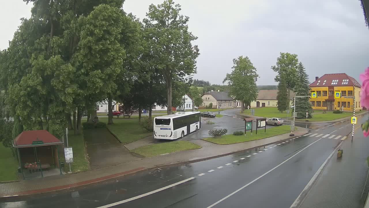 A white bus is parked in a town square on a rainy day, surrounded by buildings, trees, and a wet road.