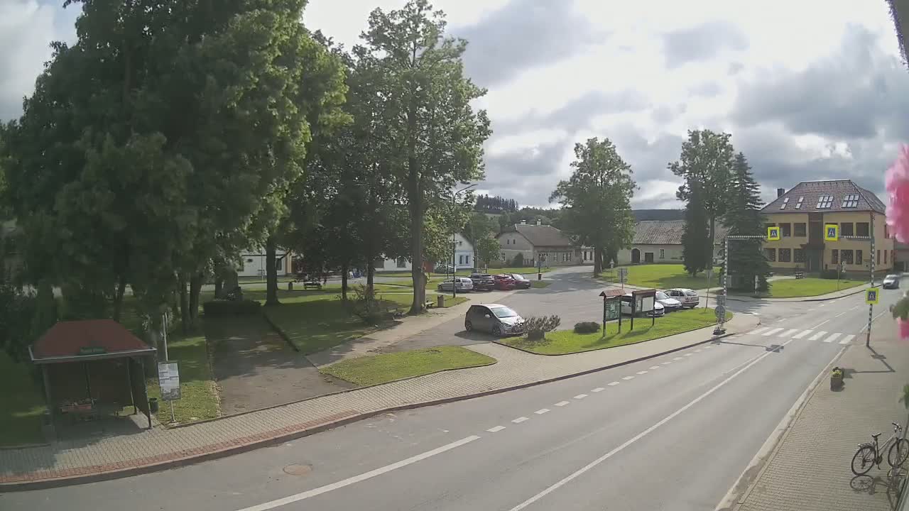 A mostly sunny day reveals a village square with several parked cars, a small park, and buildings, including a two-story structure.